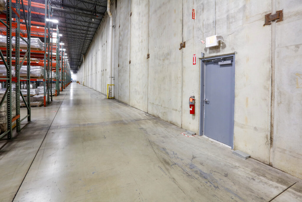 A concrete warehouse corridor with metal storage racks on the left and a gray door with an exit sign on the right.