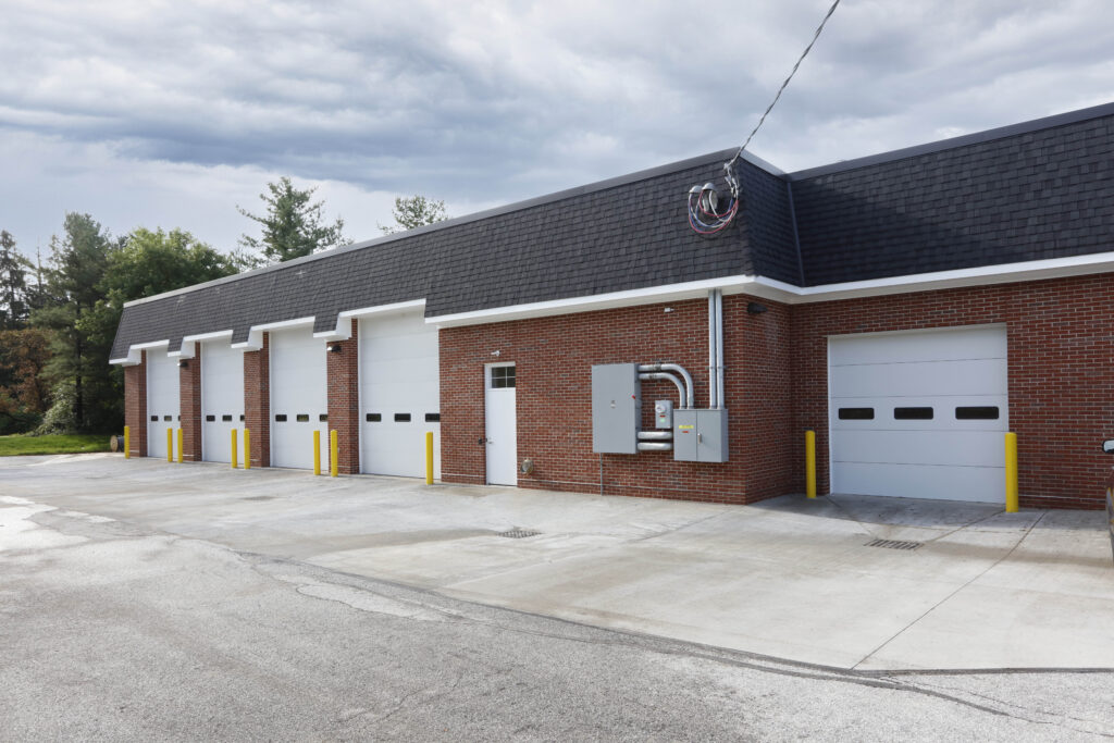 A red brick commercial building with five white garage doors, a single entry door, and yellow bollards in a paved parking lot.