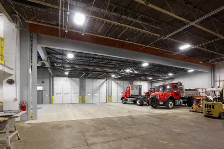 Interior of a maintenance garage with two red semi-trucks and a yellow forklift parked near three closed bay doors.