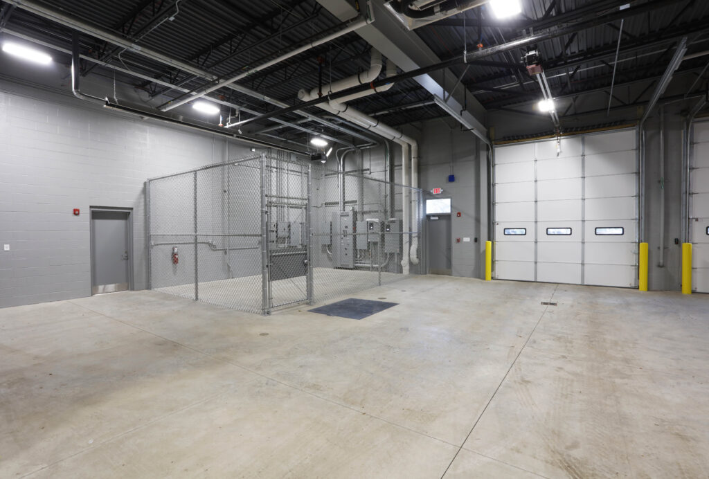 An empty warehouse space with gray walls, a chain-link storage cage, and a large roll-up metal door.