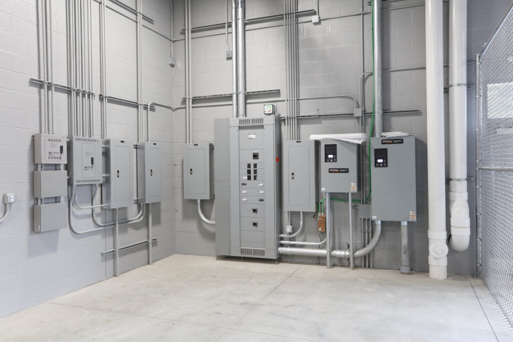 An array of gray electrical panels and conduit mounted on a concrete block wall inside an industrial room.