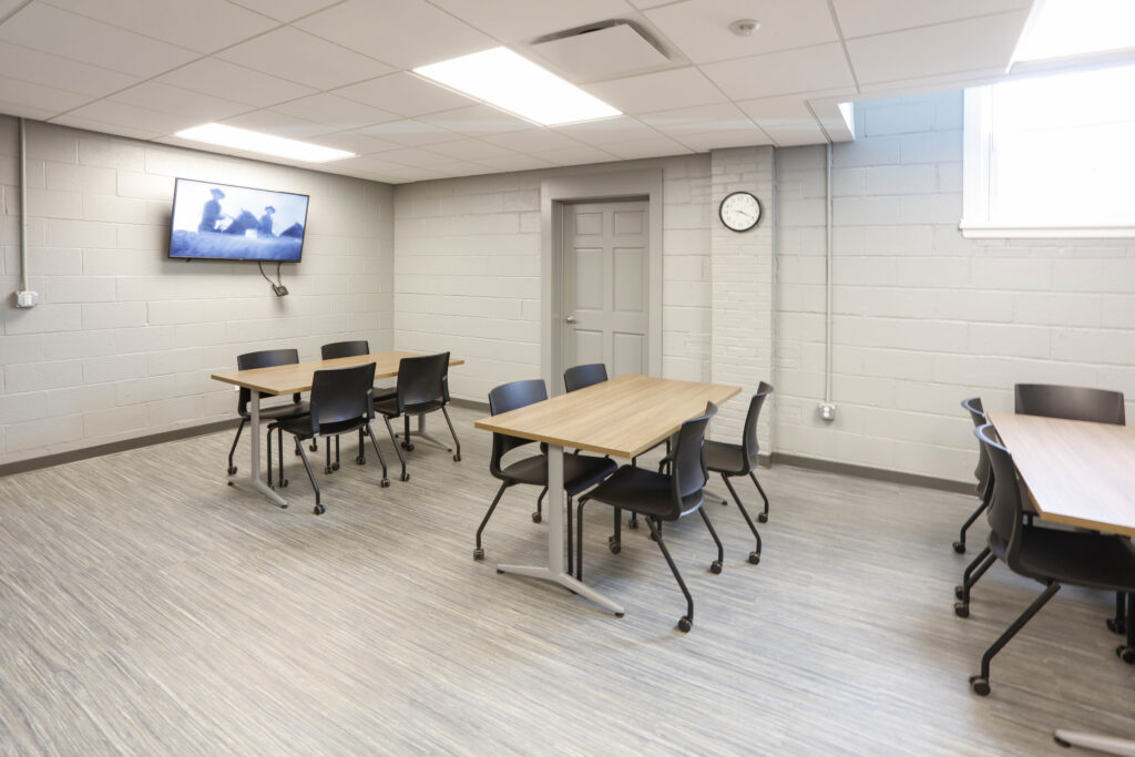 A classroom with three tables, black chairs, a wall-mounted monitor, and a clock on a cinder block wall.