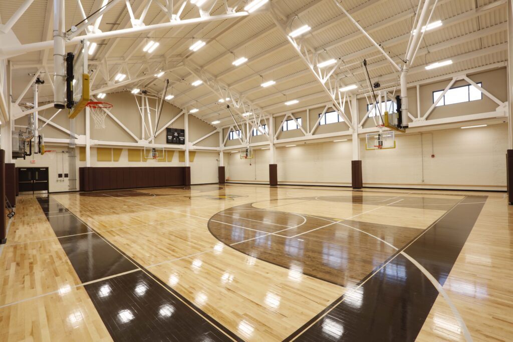 An indoor basketball court with two hoops, light-colored wood flooring, and a scoreboard on the wall.