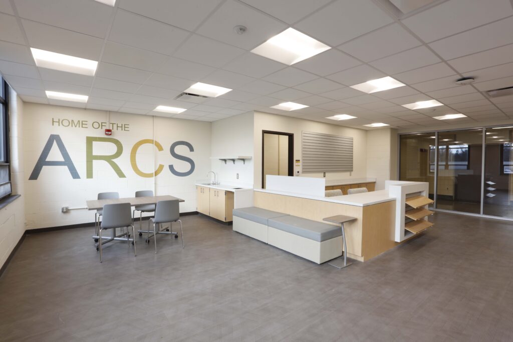 An office break room with a table and chairs, a sink cabinet, and built-in bench seating, with “HOME OF THE ARCS” printed on the wall.