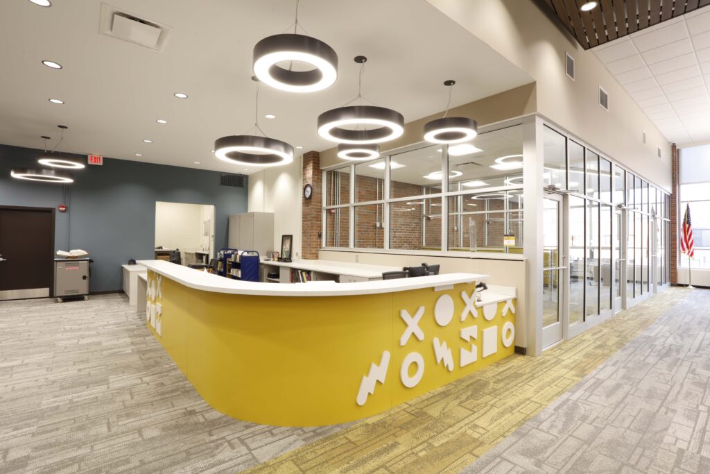 A curved yellow reception desk with geometric white shapes, situated in a brightly lit, modern office lobby with glass walls and circular pendant lights.