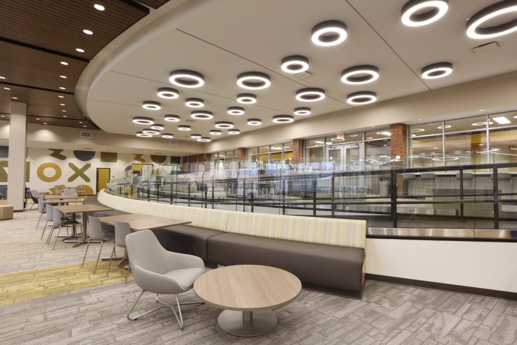 A modern library seating area with a long curved bench, small circular tables, and grey chairs under a ceiling with numerous ring-shaped lights.