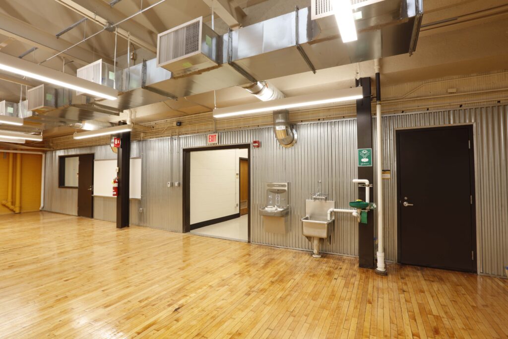 An open room with hardwood floors, a corrugated metal wall, a doorway, a water fountain, and an emergency eye wash station.