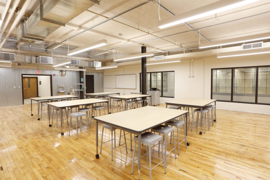 A classroom with hardwood floors featuring several light-colored rectangular tables and grey stools.