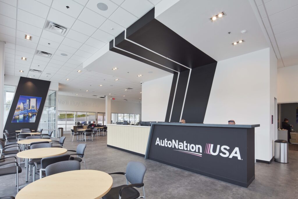 The interior of an AutoNation USA car dealership showing a reception desk, a seating area with round tables, and a wall mural of a bridge.