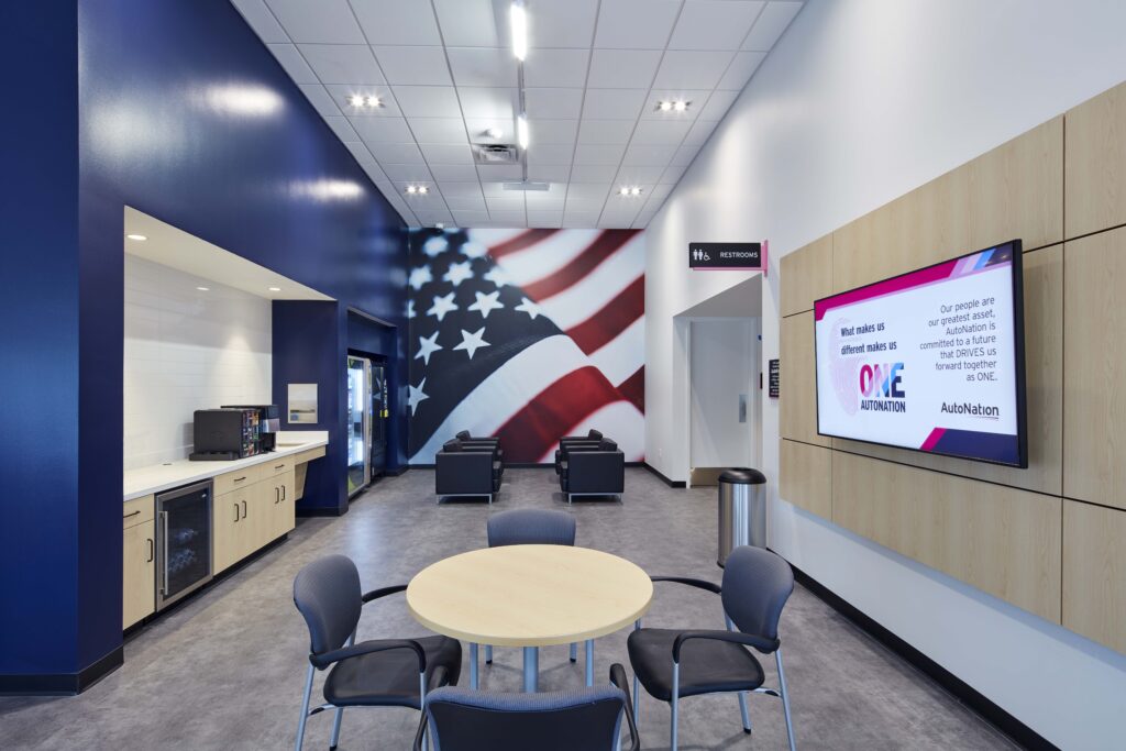 A waiting area with a round table, four chairs, a coffee station, and a wall-mounted digital screen displaying AutoNation branding, with a large American flag mural at the end of the room.