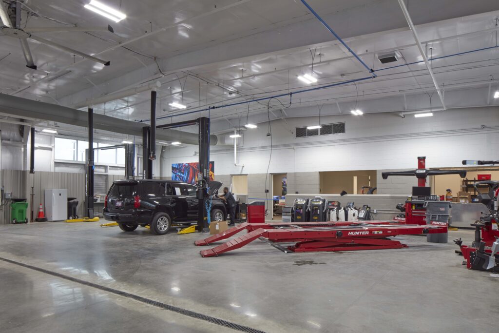 A mechanic works on a black SUV lifted in a large auto repair shop, with a red wheel alignment rack in the foreground.