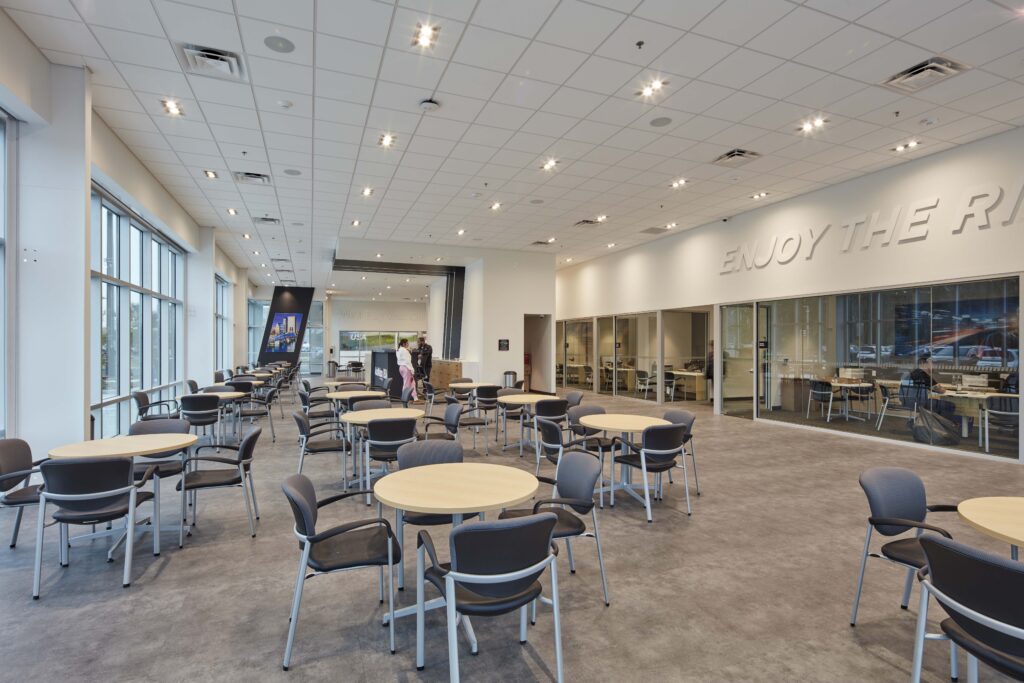 An open car dealership waiting area with several round tables, chairs, and a reception desk in the distance.