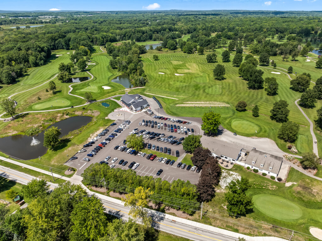 Aerial view of a golf course with a clubhouse, parking lot, ponds, and surrounding forest.