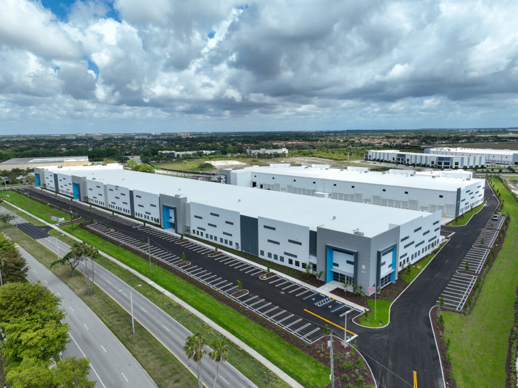 Aerial view of a large white commercial warehouse complex with blue accents, adjacent parking lots, and surrounding roads.