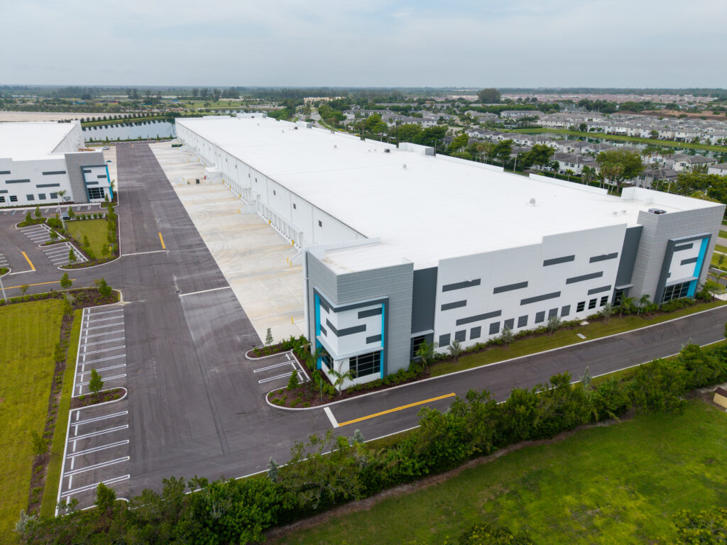 Aerial view of a large white industrial warehouse with multiple loading docks, adjacent to a parking lot and a residential neighborhood.