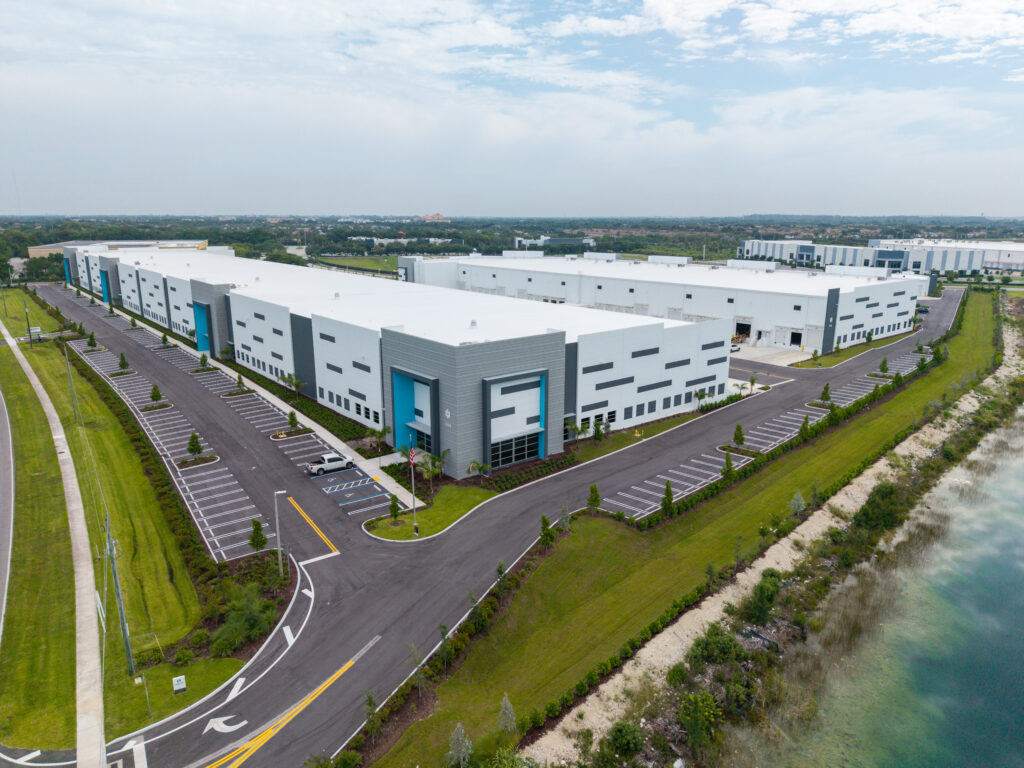 Aerial view of a large white industrial warehouse building with blue accents, surrounded by a parking lot, green spaces, and a nearby body of water.