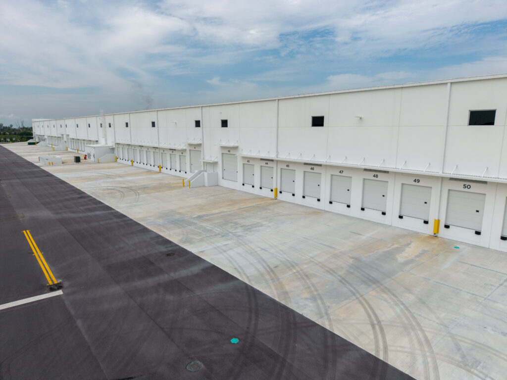 An aerial view of a long warehouse building featuring a row of numbered shipping dock doors and a paved loading area with tire tracks.