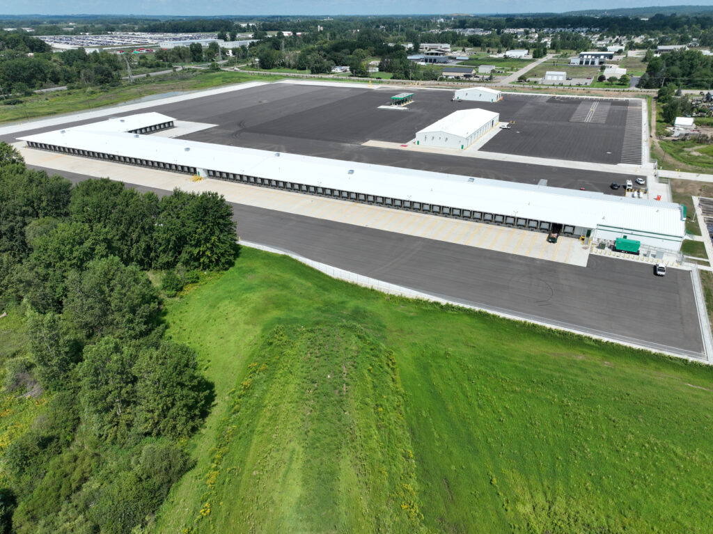Aerial view of a large warehouse complex with a long shipping bay, smaller detached buildings, and an expansive paved parking lot surrounded by grassy fields and trees.