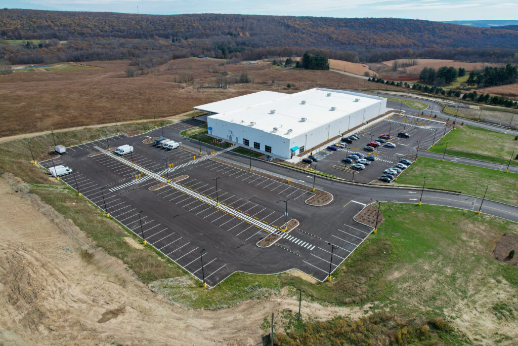 Aerial view of a white warehouse building with a large paved parking lot in a rural area.