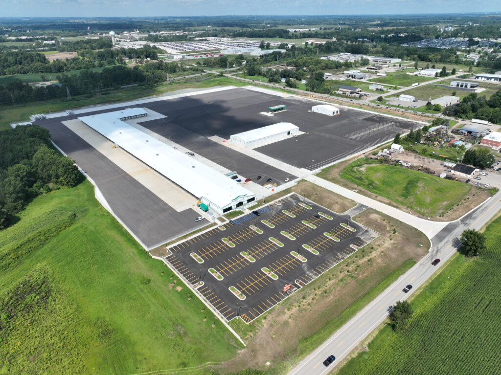 Aerial view of a long warehouse building with an adjacent large parking lot and smaller outbuildings, surrounded by rural landscape and a road.