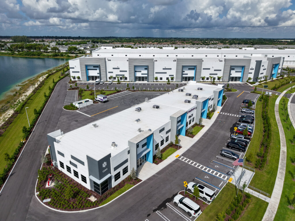 Aerial view of two adjacent modern industrial warehouse buildings with parking lots and a nearby lake.