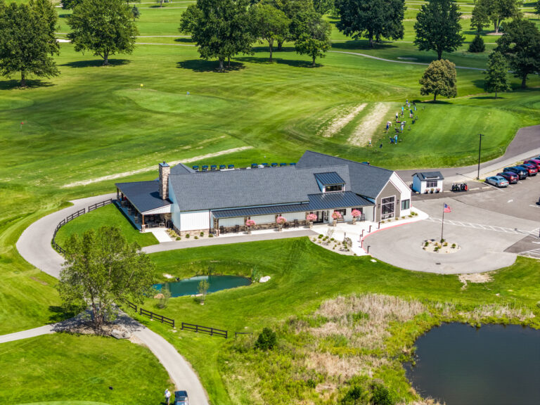 Aerial view of a golf course clubhouse with a parking lot, a pond, and golfers walking on the fairway.