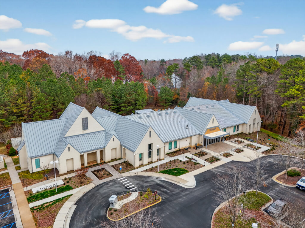 Aerial view of a multi-winged, light-colored building with a grey metal roof, surrounded by trees and a paved parking lot.
