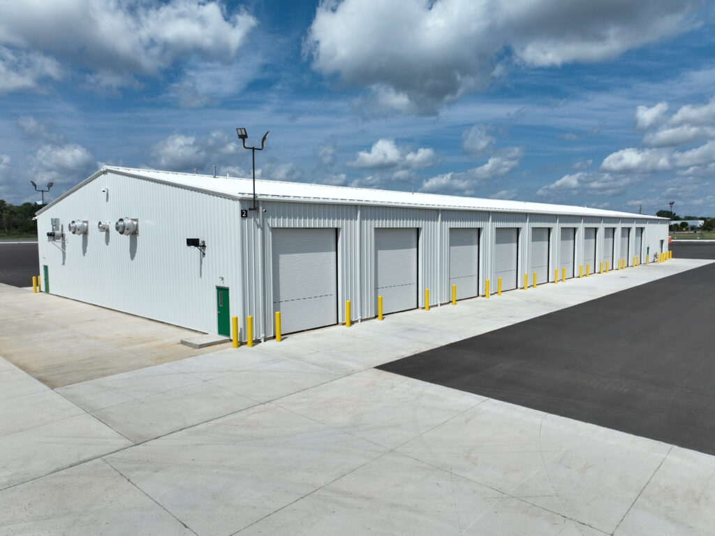 A white industrial warehouse building with multiple roll-up doors and yellow safety bollards.