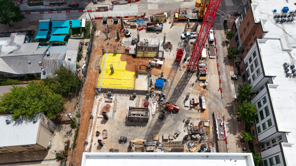Aerial view of a construction site featuring a red crane, foundation work with yellow protective covers, and scattered building materials.