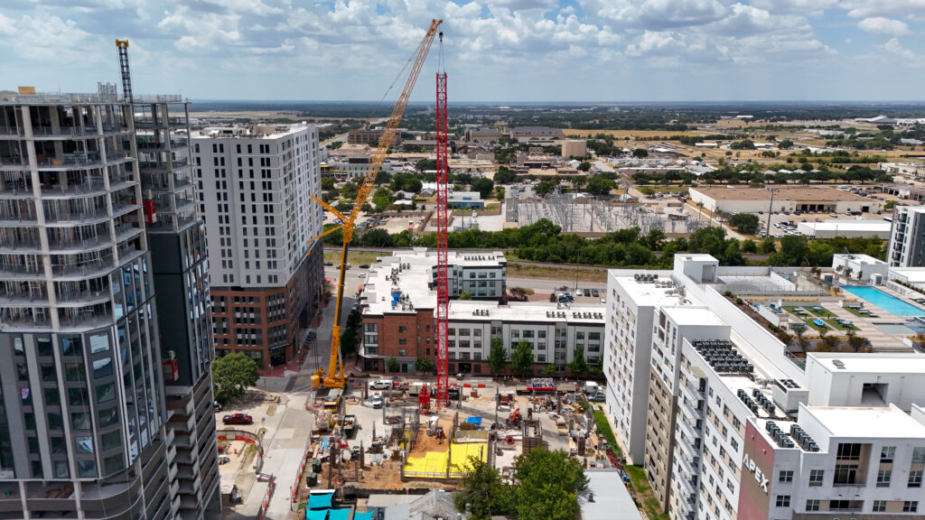 Aerial view of a construction site featuring a tall red crane and a yellow mobile crane between several modern apartment buildings.