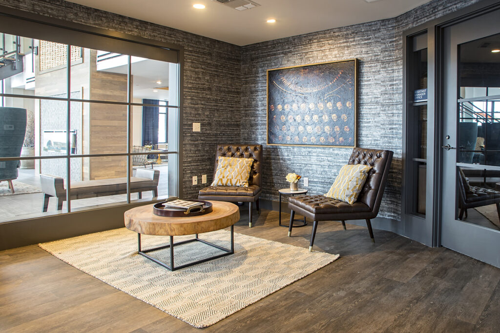 Two brown leather armchairs with patterned pillows, a round wooden coffee table, and framed artwork in a lobby waiting area.