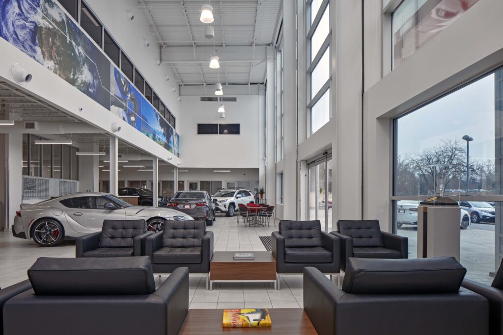 Modern car dealership showroom with black leather lounge seating in the foreground and various cars displayed throughout the space.