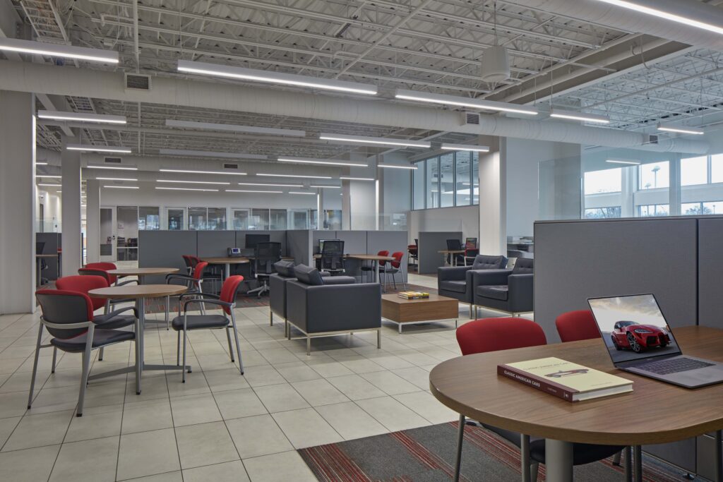 Modern open-plan office with cubicles, tables, chairs, and a laptop displaying a red sports car.