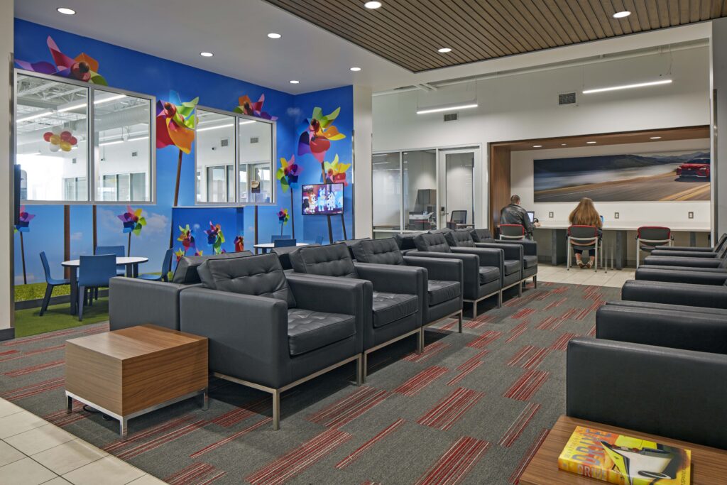A waiting area with rows of black leather armchairs, a wall decorated with colorful pinwheels, and a workspace with two people using computers.