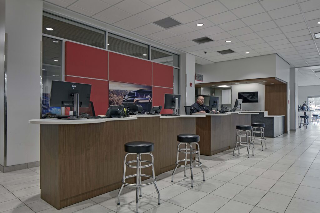 A service counter in a car dealership featuring wood panels, black stools, computer terminals, and a staff member sitting at the desk.