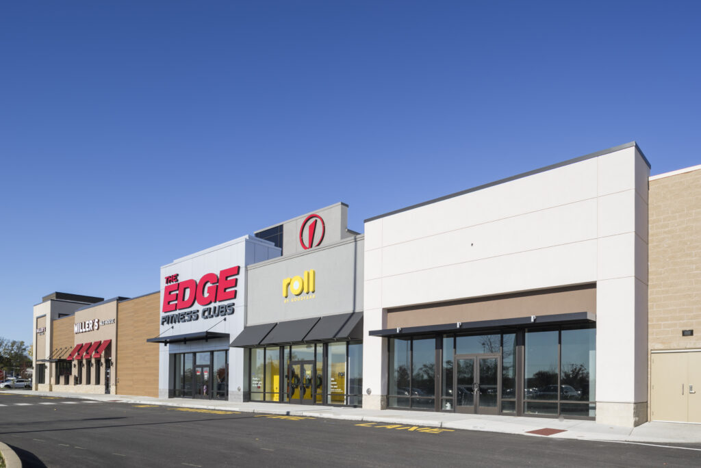 The exterior of a strip mall featuring The Edge Fitness Clubs, Roll by Goodyear, and other storefronts under a clear blue sky.