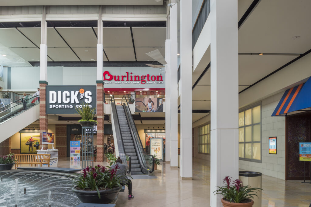 Interior of a multi-level shopping mall featuring an escalator, a Burlington store, a Dick’s Sporting Goods store, and a fountain in the foreground.