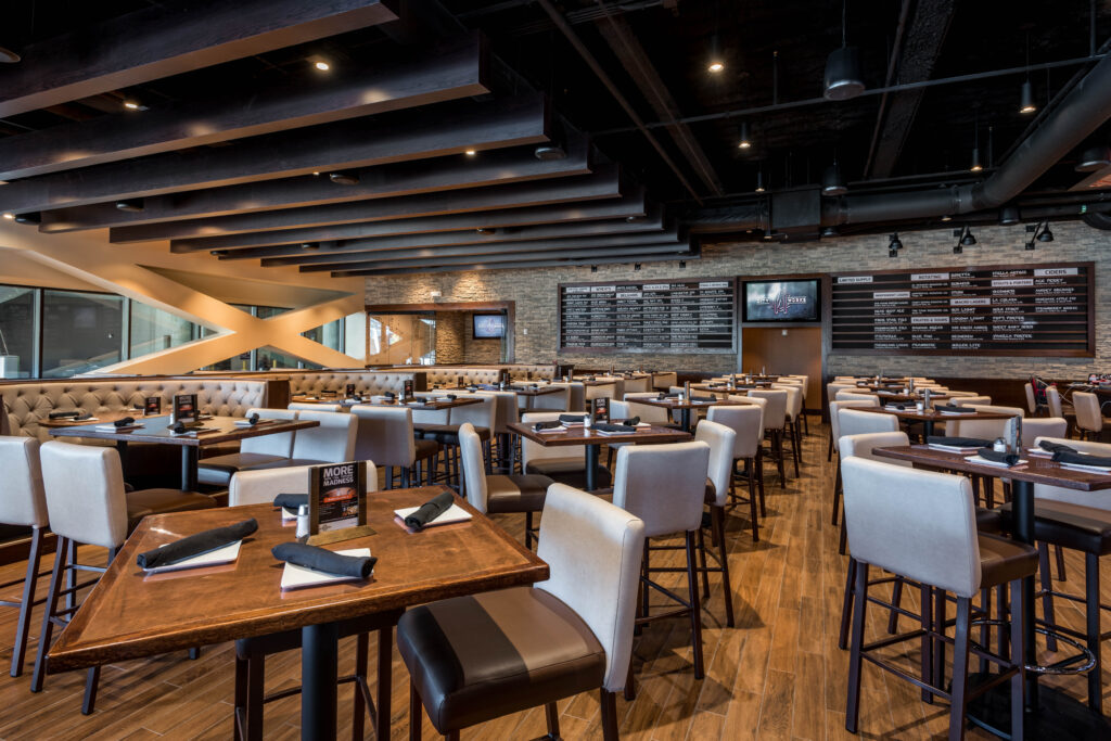 The interior of a restaurant with rows of tables and chairs, wood flooring, and large menu boards displaying beer selections on the wall.