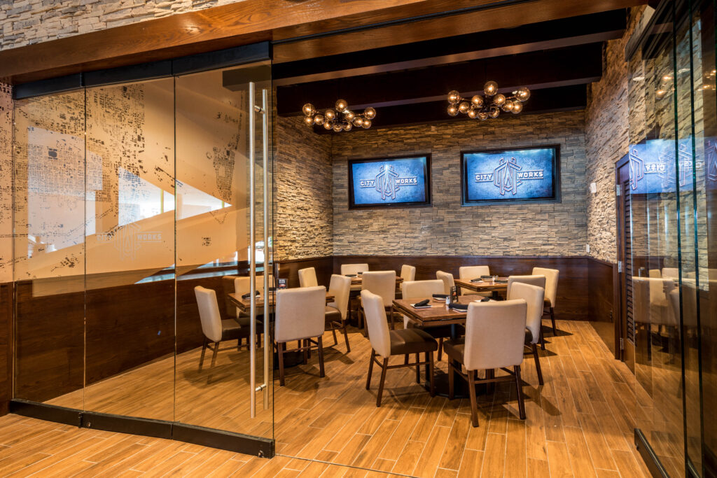 Interior of a City Works restaurant featuring dining tables with chairs, stone walls, and two screens displaying the City Works logo.