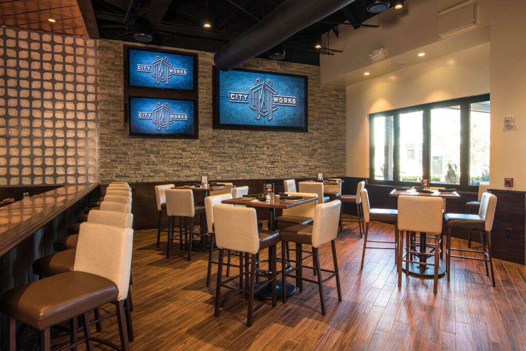 The interior of a restaurant featuring wooden floors, high-top tables, and screens displaying the City Works logo on a stone wall.