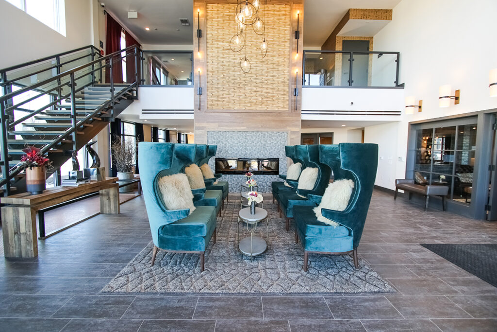 Lobby seating area with two rows of teal high-back chairs facing a central fireplace and a floating staircase.