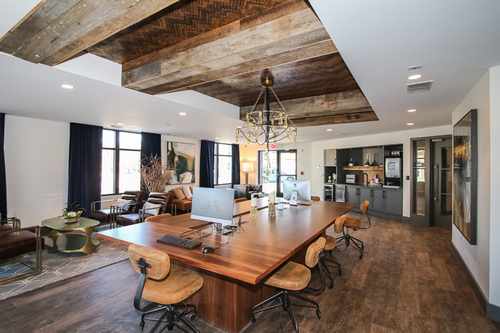 Modern coworking space featuring a long wooden conference table with desktop computers, surrounded by office chairs, with a kitchenette and lounge area in the background.