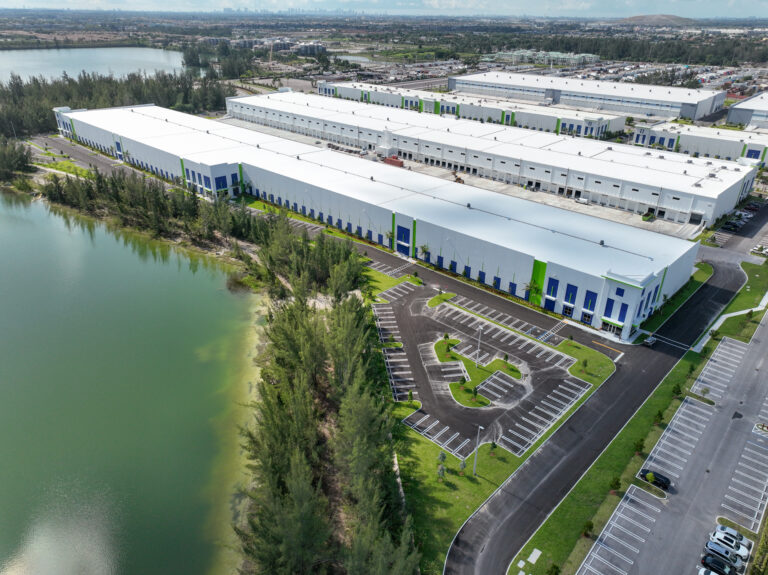 Aerial view of large white warehouse buildings situated next to a lake and parking lots.