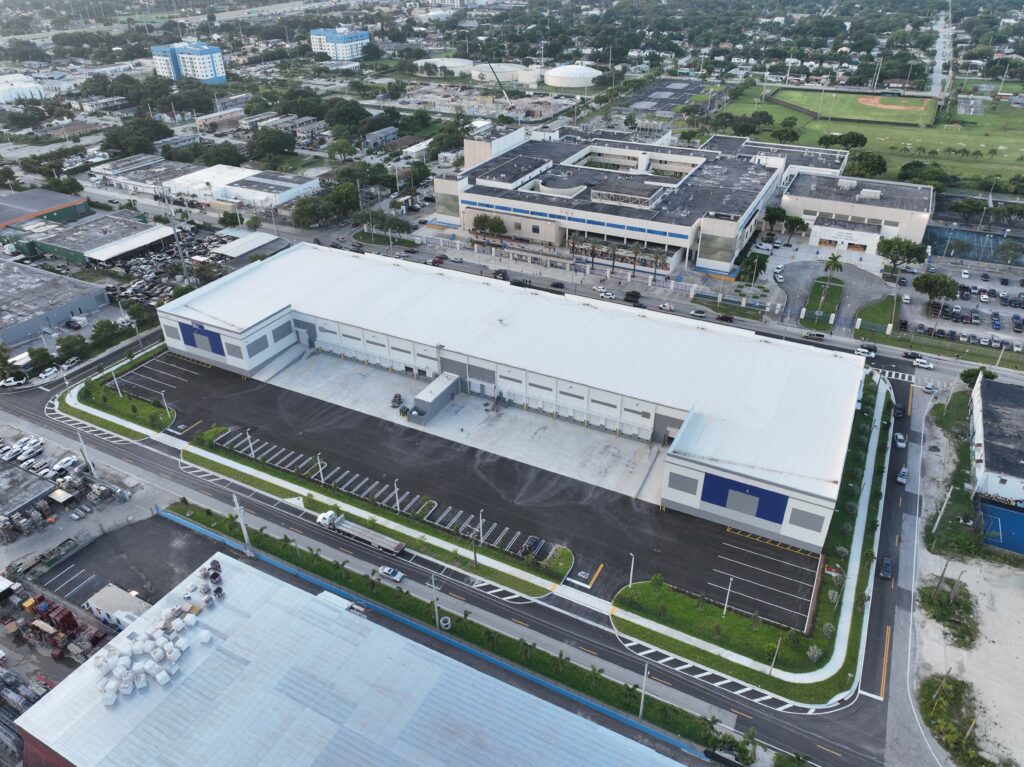 Aerial view of a large rectangular warehouse with loading docks, adjacent parking, and a nearby school building.