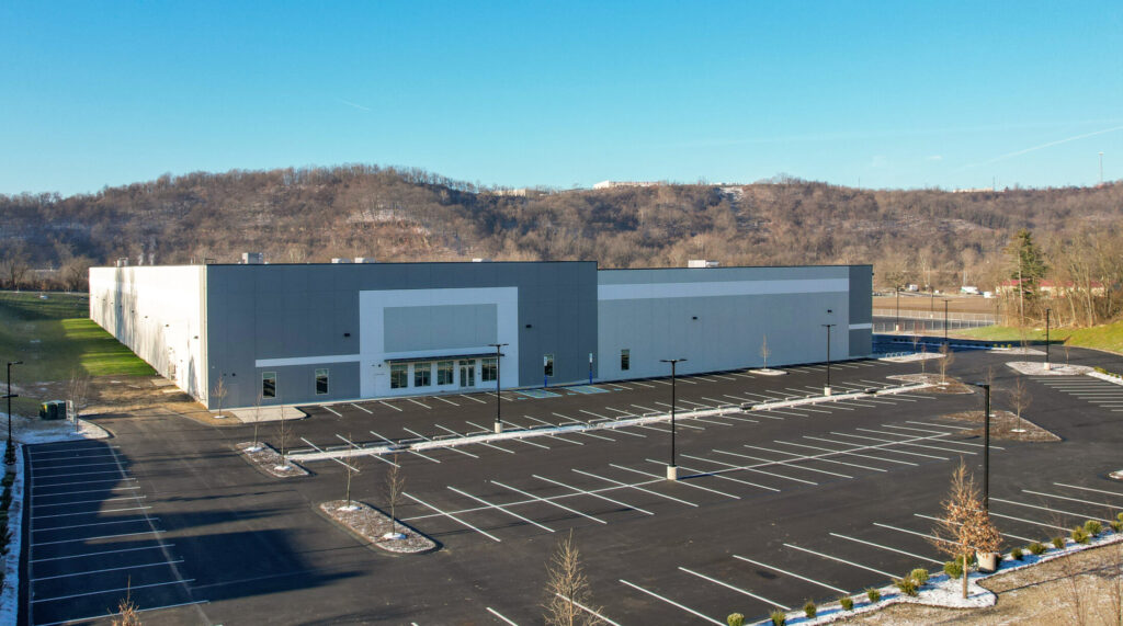 An aerial view of a large, gray warehouse building with an empty parking lot in the foreground, set against a hilly landscape.