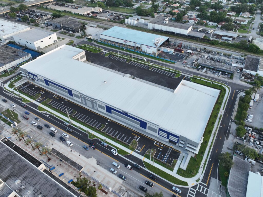 Aerial view of a large rectangular warehouse building with a white roof and blue accents, featuring an adjacent parking lot and surrounding industrial area.