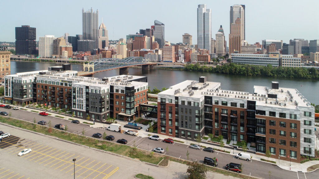 Aerial view of an apartment complex with a parking lot in the foreground and the Pittsburgh skyline and a bridge across the river in the background.