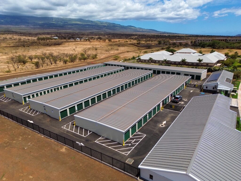 Aerial view of a self-storage facility with multiple long, gray-roofed buildings and parking spaces, situated in an arid landscape.