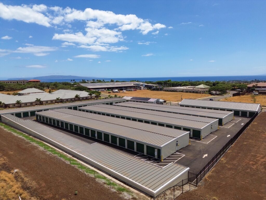 Aerial view of a self-storage facility with multiple rows of long metal buildings and green roll-up doors, with the ocean in the distance.