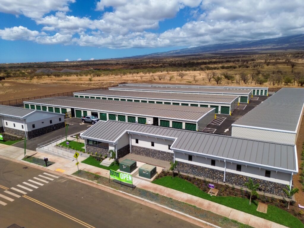 Aerial view of a self-storage facility with multiple rows of units and a front office building displaying a “Now Open” sign.